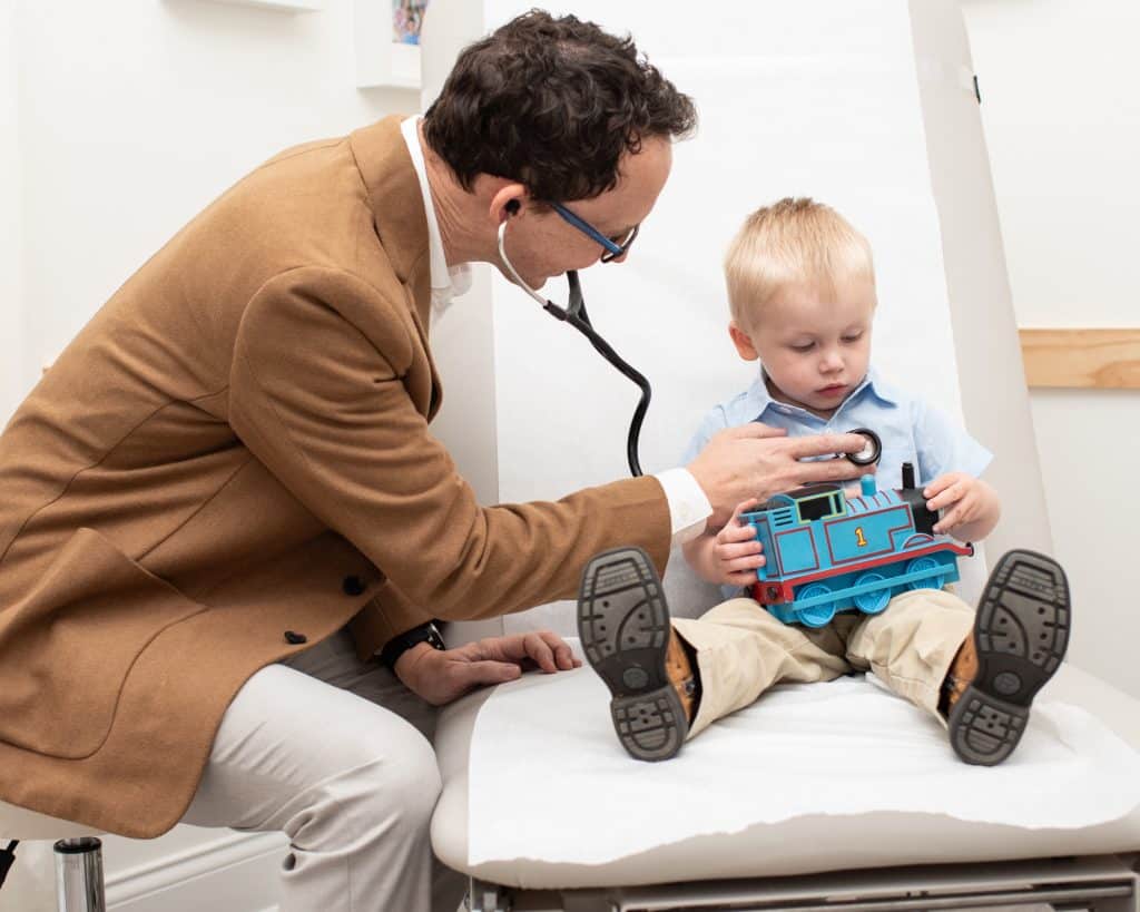 Doctor checking the heart of a small male child holding a Thomas the Tank engine.